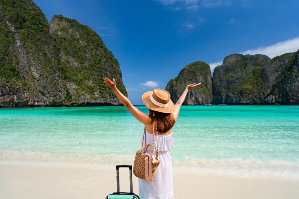 Goedkoop vliegen naar een tropisch eiland met een reiziger die blij op het strand staat met haar koffer.